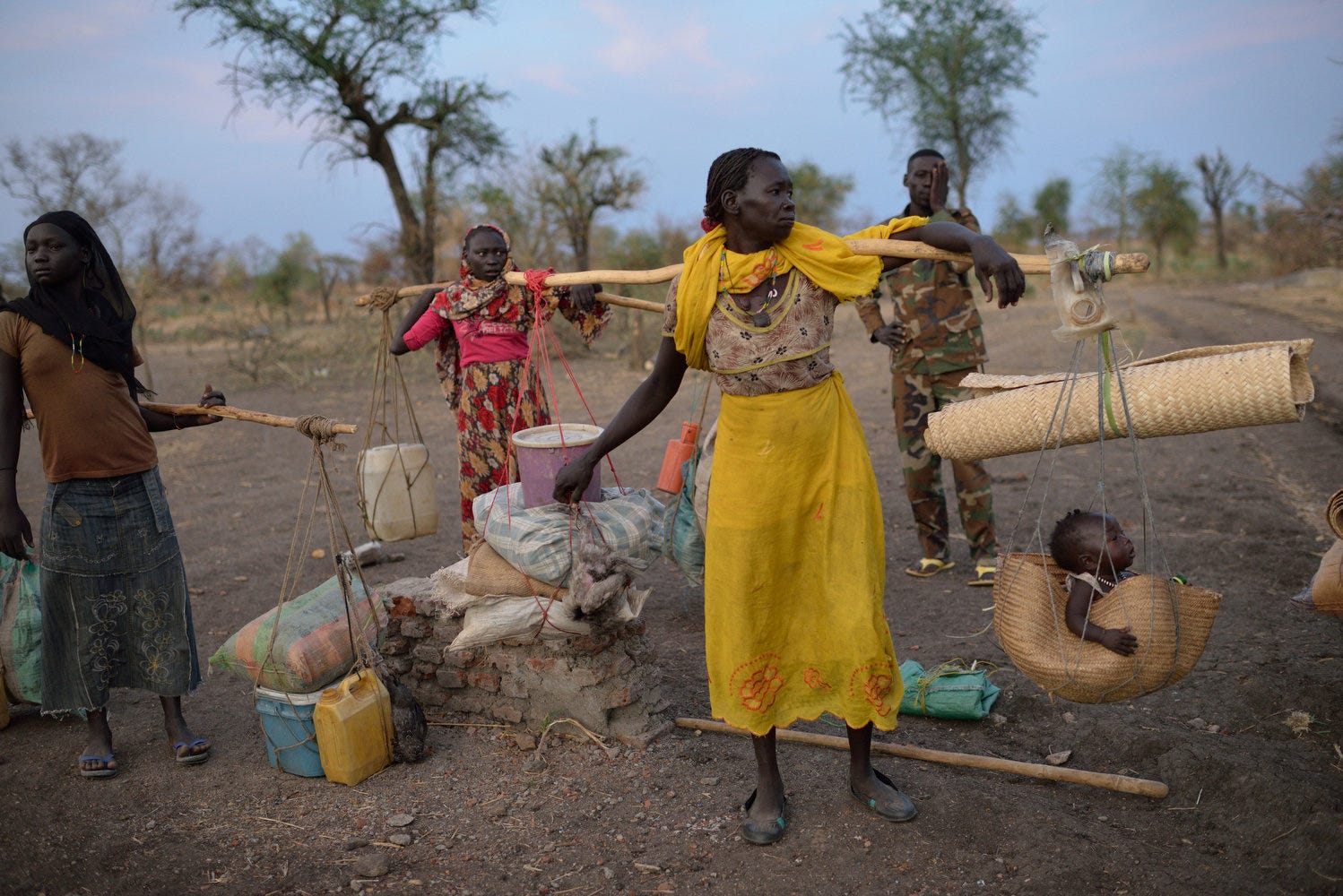 four people carry belongings about their shoulders on yokes. they wear open twoed shoes. Two persons are in hijabs covering their hair in neck. the person most in the foregrand is wearing bright yellow wrapped around their waist, and has a baby fashioned in a basket hanging off the yoke. 